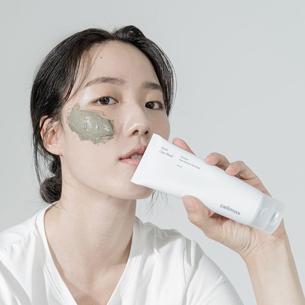 Una donna applica una maschera all'argilla verde, tenendo un tubo CELIMAX "The Real Noni Refresh Clay Mask".