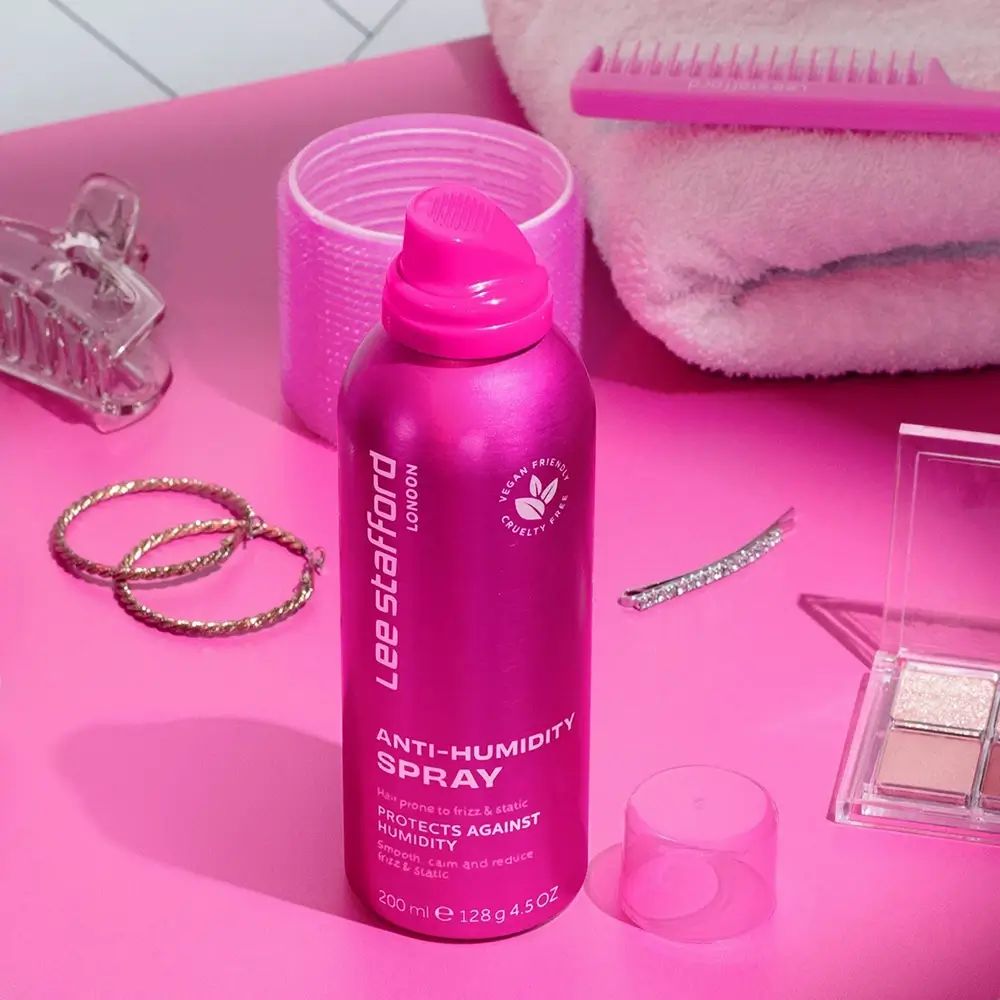 Flacone spray rosa Lee Stafford Anti-Humidity. Vegano, cruelty-free. Su sfondo rosa con accessori.