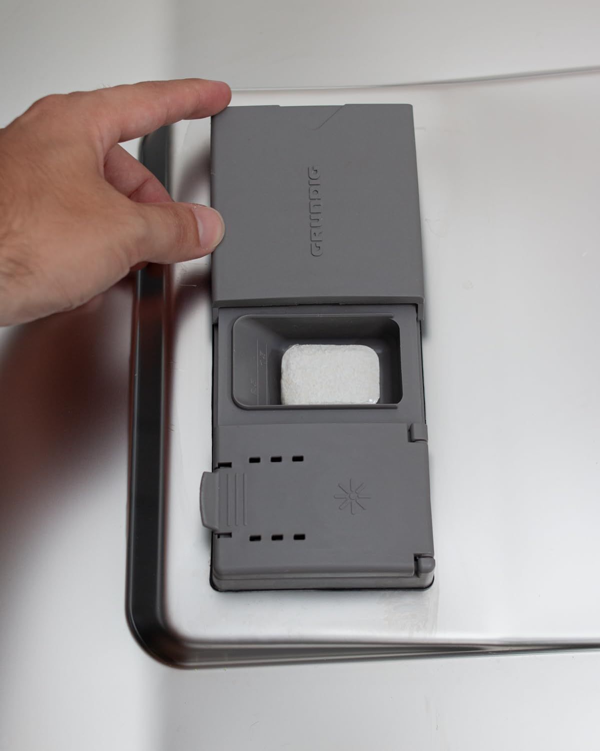 Mano apre il vano detersivo grigio di una lavastoviglie. Una pastiglia nel vano.