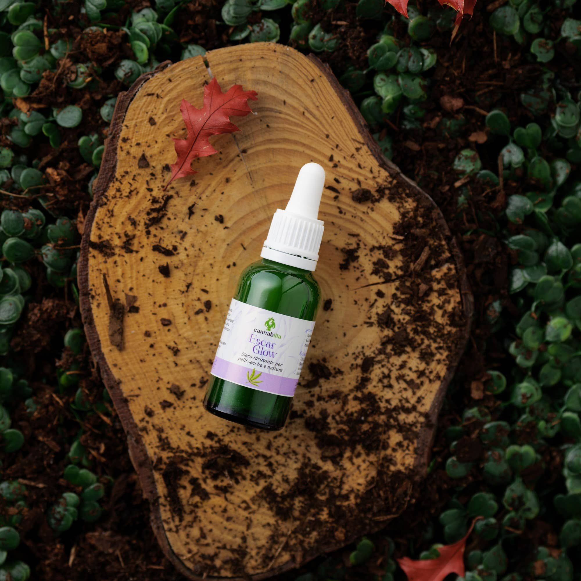 Flacone di vetro verde con contagocce bianco su un ceppo. Scritta: Escar Glow. Logo Cannabilla. Sfondo: foglie e una foglia rossa.