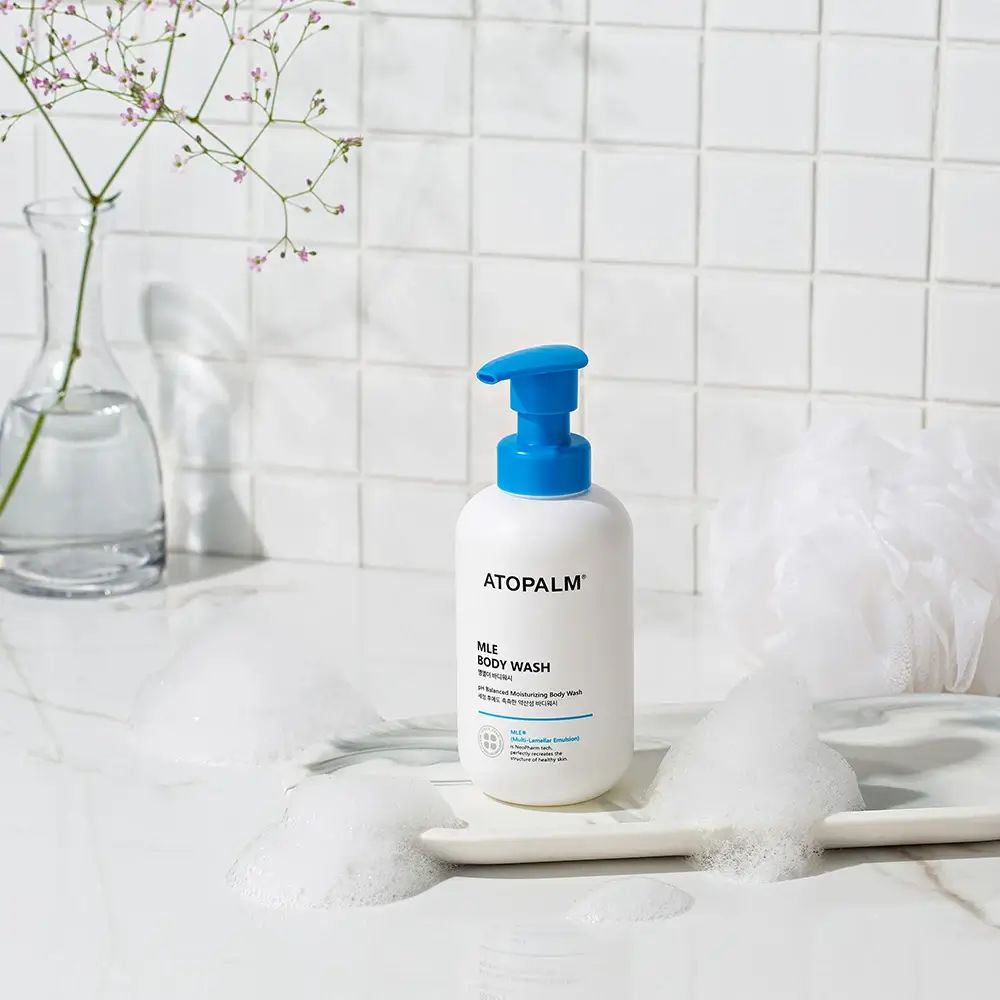 Flacone bianco con erogatore blu. Scritta: ATOPALM MLE Body Wash. Schiuma e decorazioni.