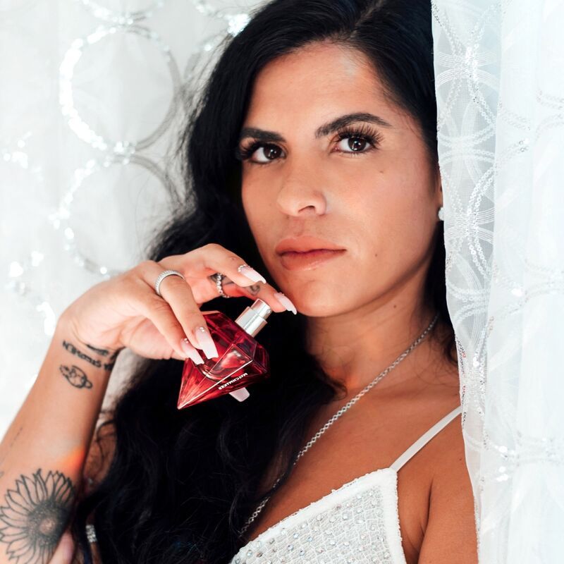 Donna che tiene in mano un flacone di profumo a forma di diamante rosso. Indossa un top bianco e ha lunghi capelli scuri.