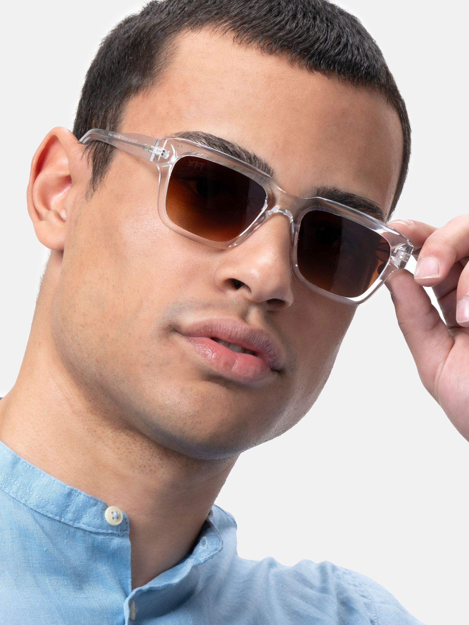Uomo con occhiali da sole trasparenti e lenti marroni. Primo piano. Camicia chiara. Mano tocca gli occhiali.