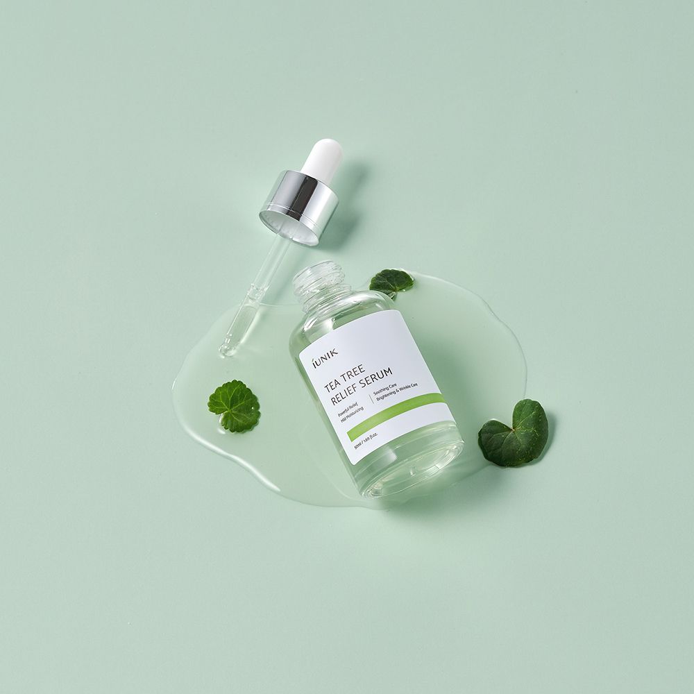 Flacone iUNIK Tea Tree Relief Serum con contagocce. Liquido su superficie con foglie. Sfondo verde chiaro.