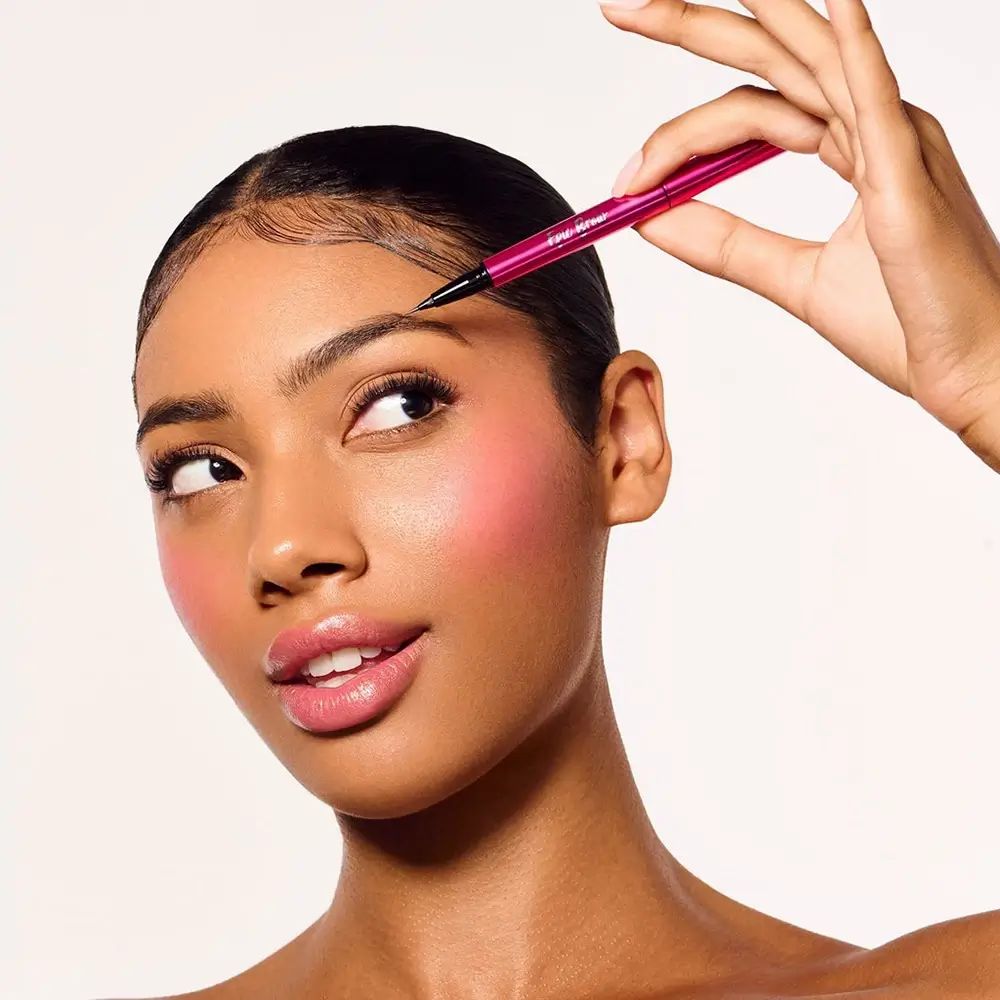Una persona tiene in mano una matita per sopracciglia rosa. La matita è posizionata sulla sopracciglia. Il prodotto è etichettato « Epic Brow ».