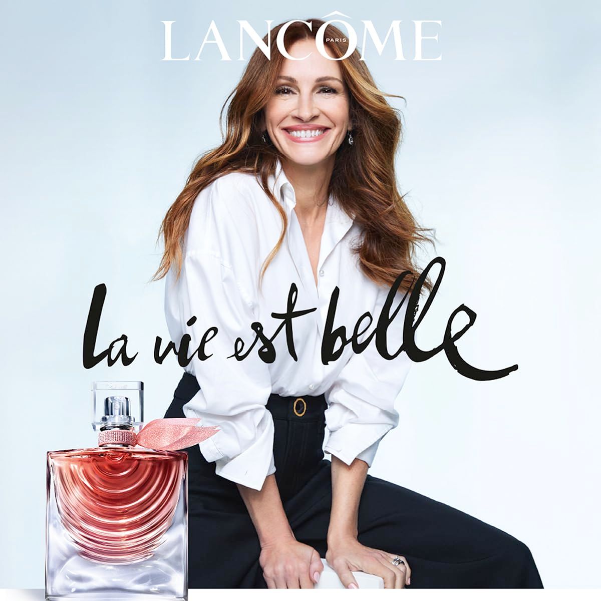 Donna con camicia bianca e pantaloni neri. Flacone in vetro con liquido rosa. Testo: Lancôme, La vie est belle.