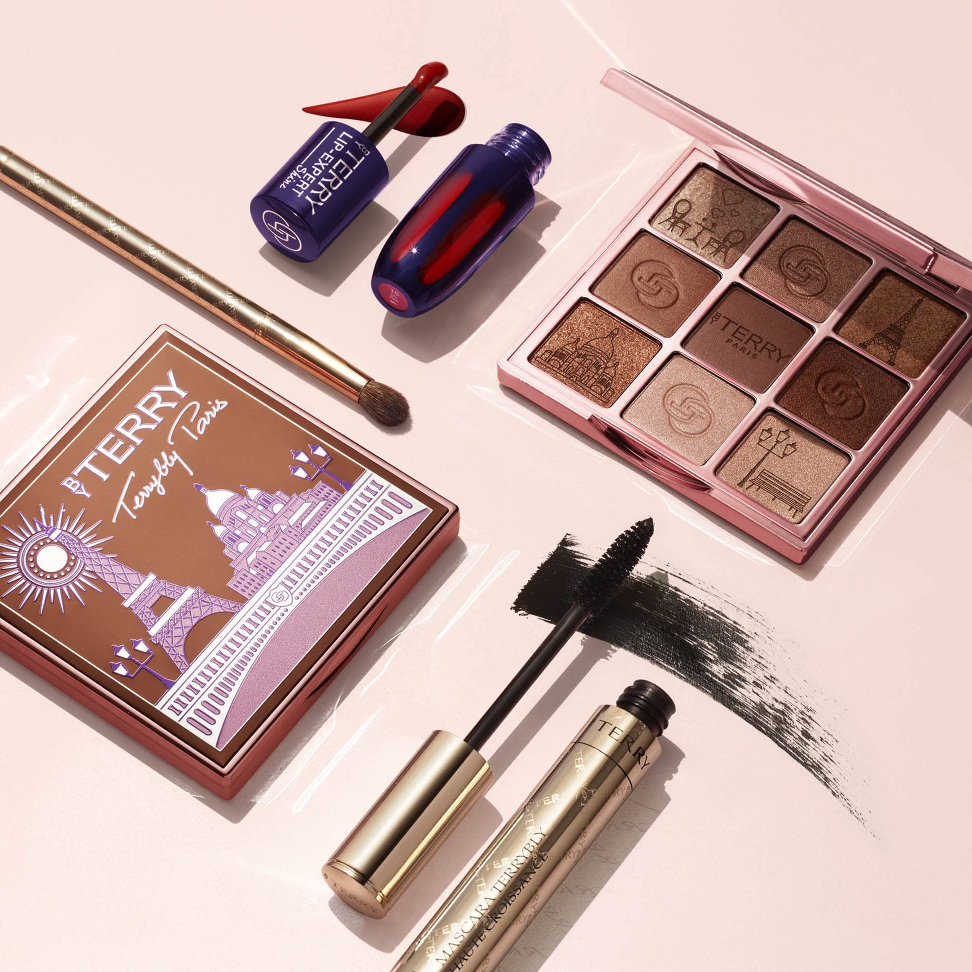 Prodotti per il trucco su superficie rosa. Mascara, palette ombretti, rossetto, pennello. Testo: Terry.
