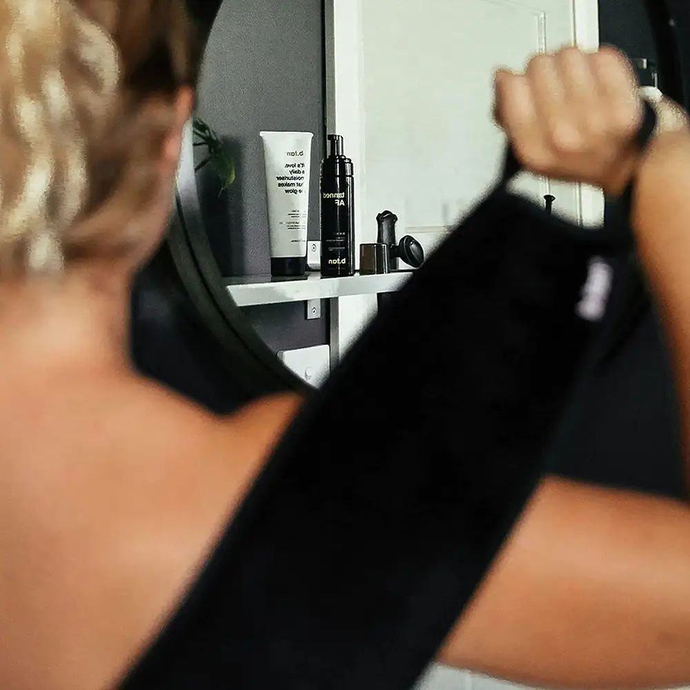 Persona che tiene in mano un applicatore per abbronzatura posteriore nero. Specchio sullo sfondo, mensola con prodotti.