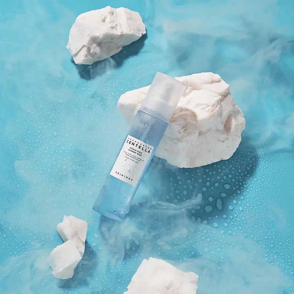 Flacone spray blu trasparente su sfondo blu con pietre bianche e nebbia. Scritta: MADAGASCAR CENTELLA, SKIN1004.