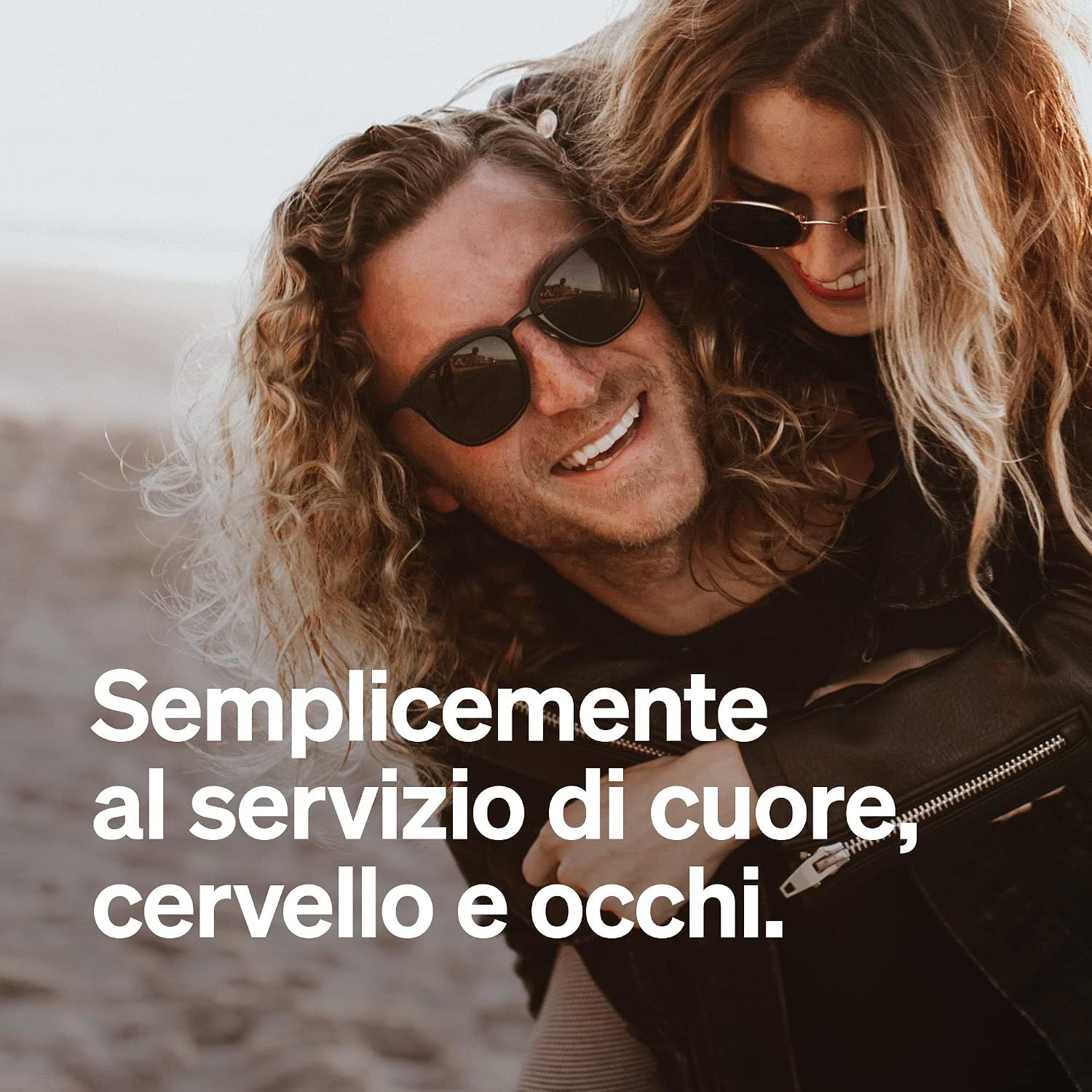 Una coppia ride e si abbraccia sulla spiaggia. Indossano occhiali da sole.