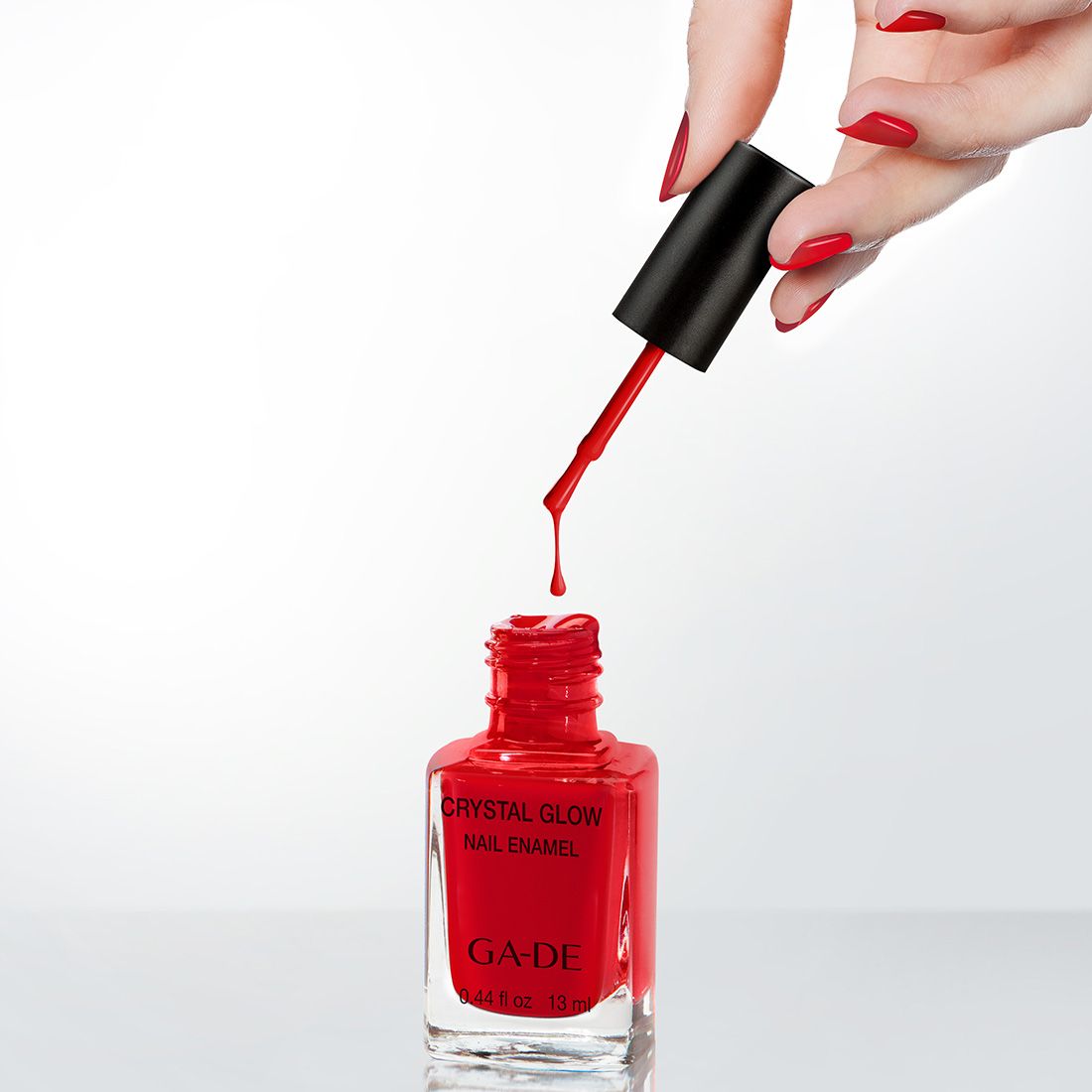 Smalto per unghie rosso in bottiglia di vetro, pennello con goccia. Scritta: Crystal Glow Nail Enamel, GA-DE. Mano con unghie smaltate.