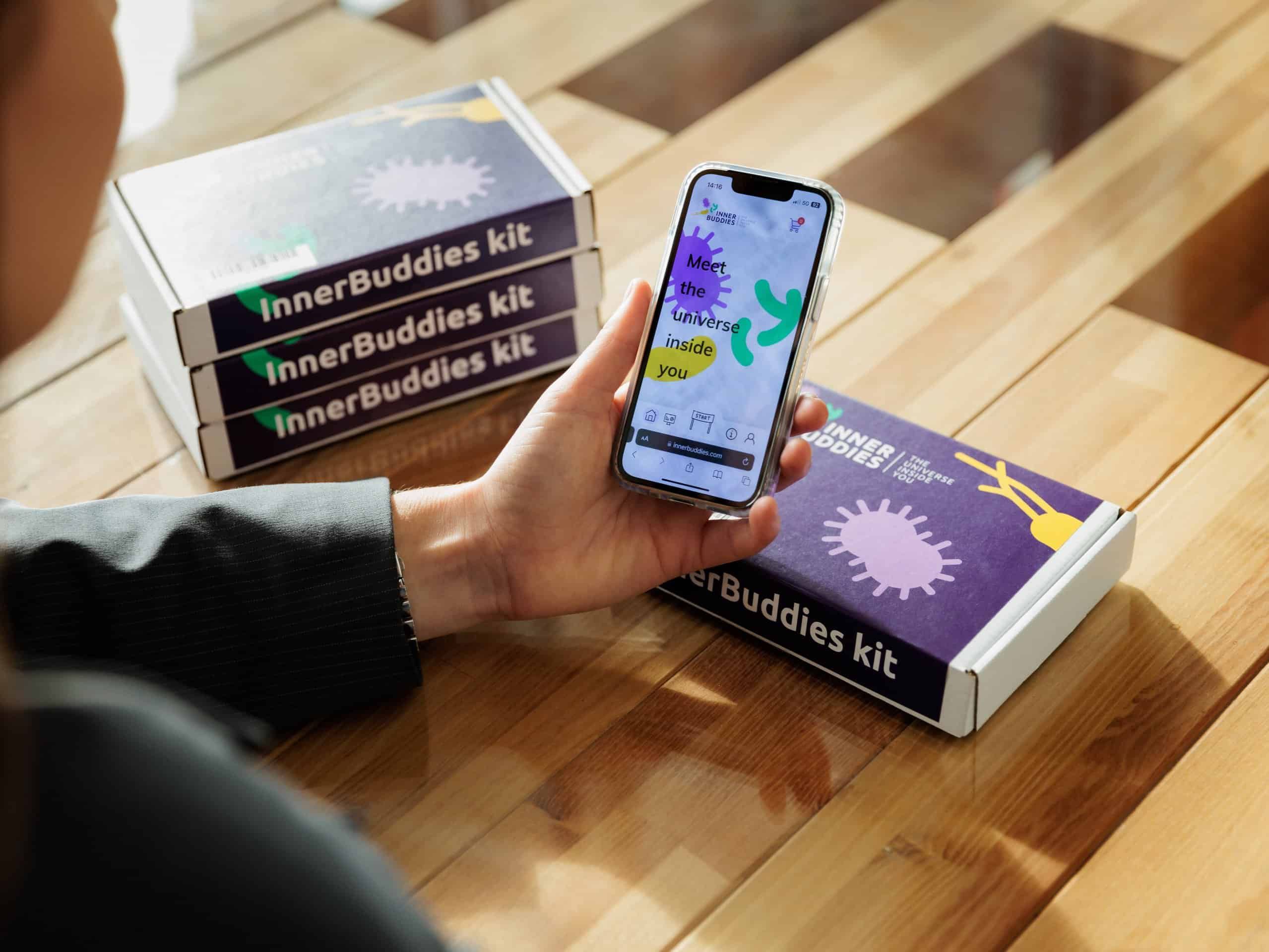 Tre confezioni di kit di test Inner Buddies impilate accanto a una persona che tiene uno smartphone.