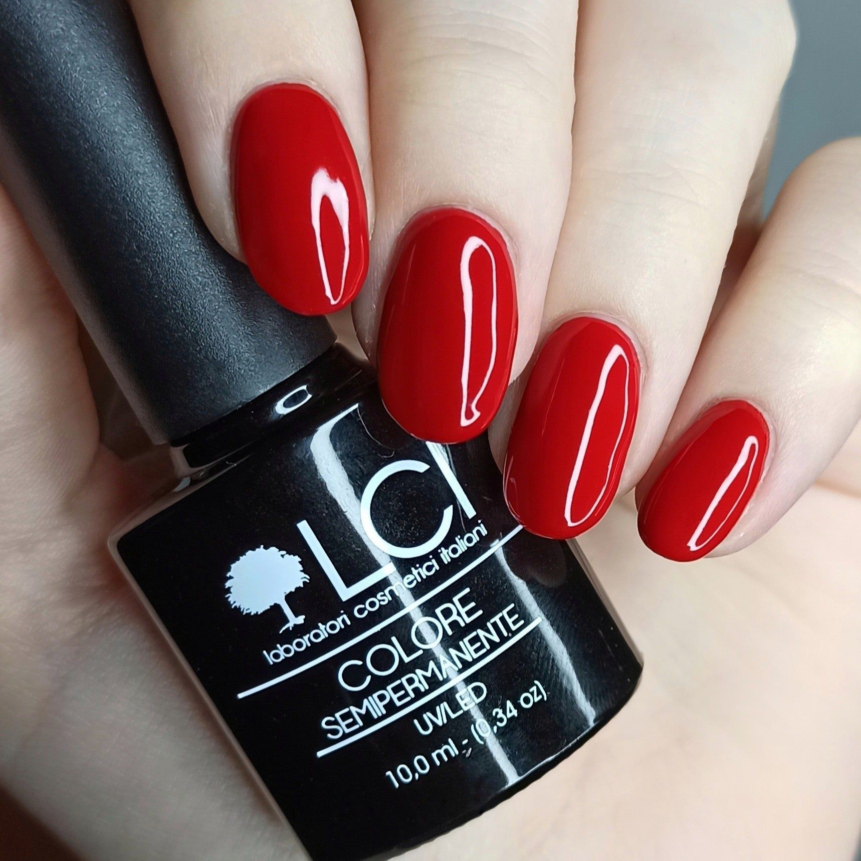 Mano con unghie rosse smaltate e flacone smalto. Logo LCI e testo: Colore Semipermanente, UVLED 10,0 ml.
