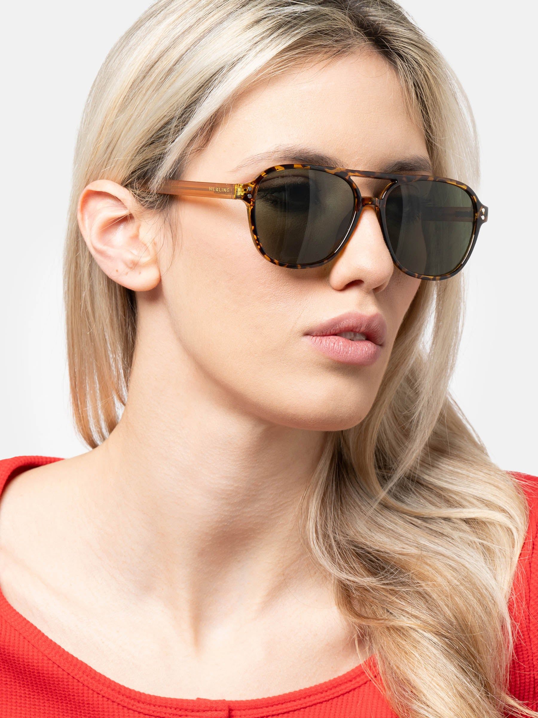 Una donna indossa occhiali da sole con montatura marrone e lenti verdi. La montatura presenta un motivo tartarugato. Il nome del marchio è visibile.