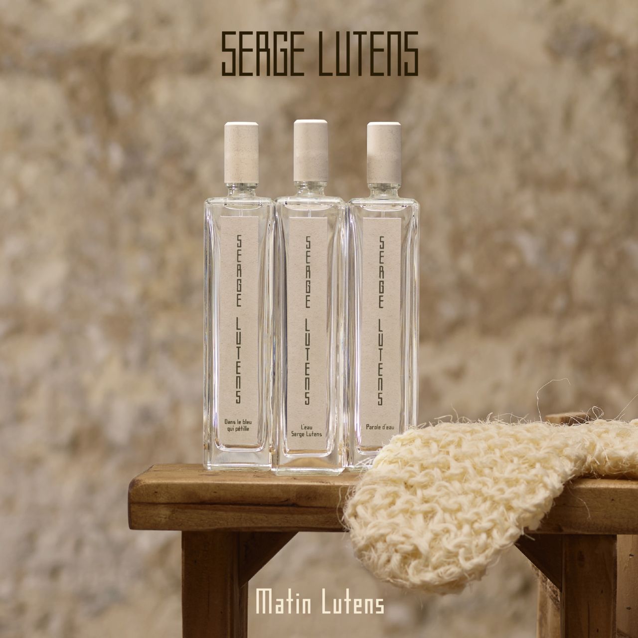Tre flaconi rettangolari in vetro su uno sgabello di legno. Scritta: SERGE LUTENS. Etichette beige, tappi bianchi.