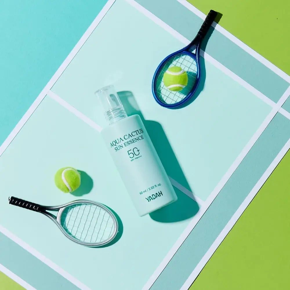 Flacone verde chiaro con applicatore bianco. Scritta: Aqua Cactus Sun Essence 50. Due palline e racchette da tennis accanto.