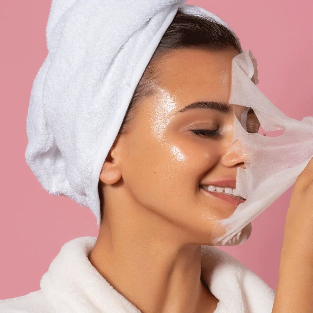 Donna con asciugamano e accappatoio che rimuove una maschera in tessuto dal viso. Sfondo rosa.