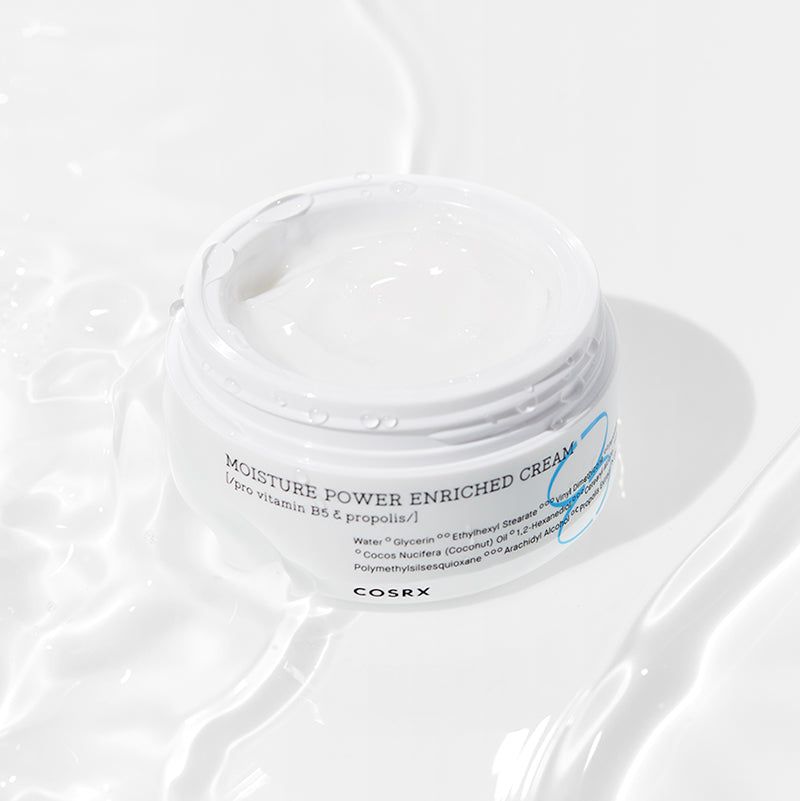 Vaso di crema aperto con superficie di crema. Scritta: MOISTURE POWER ENRICHED CREAM, COSRX. Acqua sullo sfondo.