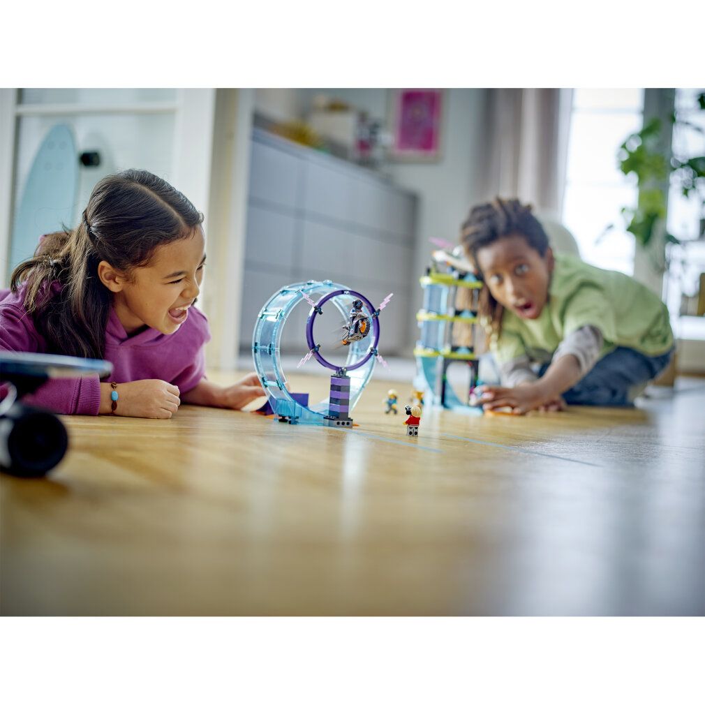 Due bambini giocano con il set LEGO City Stuntz su pavimento in legno. Pista con loop e personaggi costruiti.