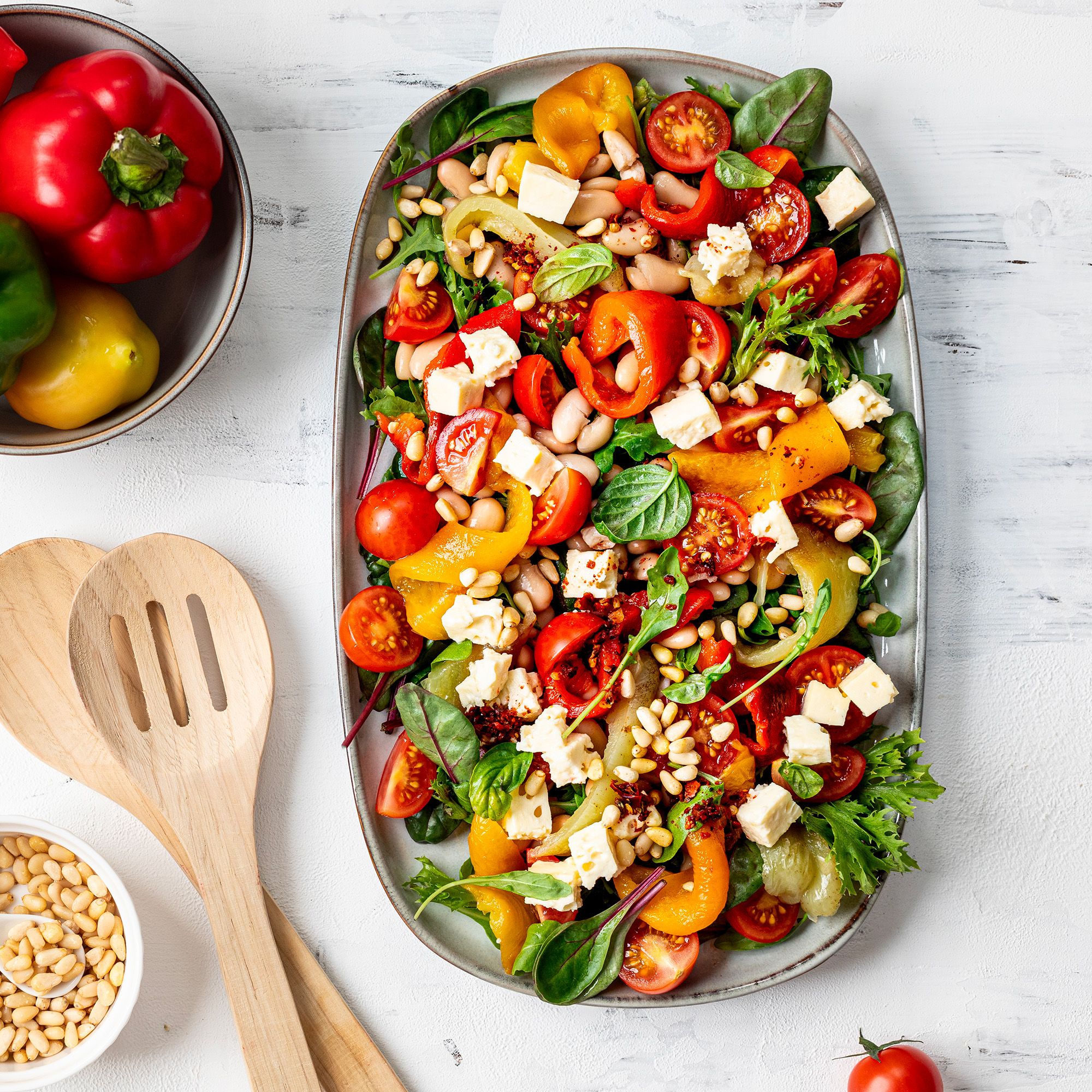 Insalata colorata con pomodori, peperoni, fagioli bianchi, feta e pinoli su un piatto ovale.