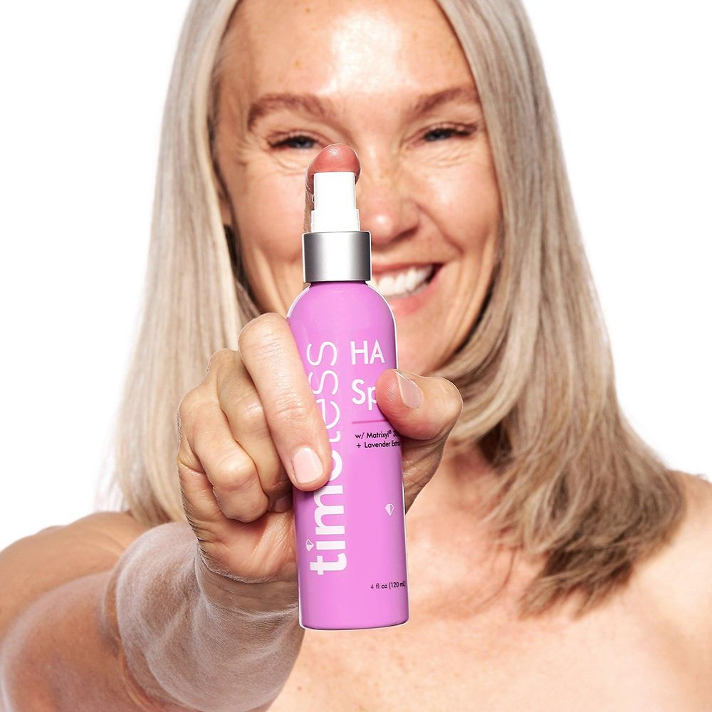Donna tiene flacone spray rosa con scritte bianche: timeless, HA Spray, Matrixyl 3000, Estratto di lavanda. Erogatore argento.