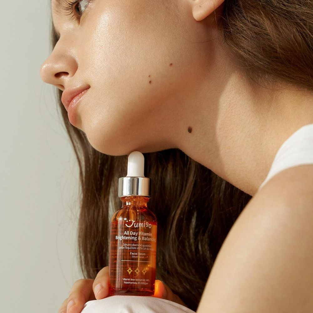 Donna che tiene in mano un flacone di siero. Scritta: JUMISO All Day Vitamin Brightening & Balancing. Viso di profilo.