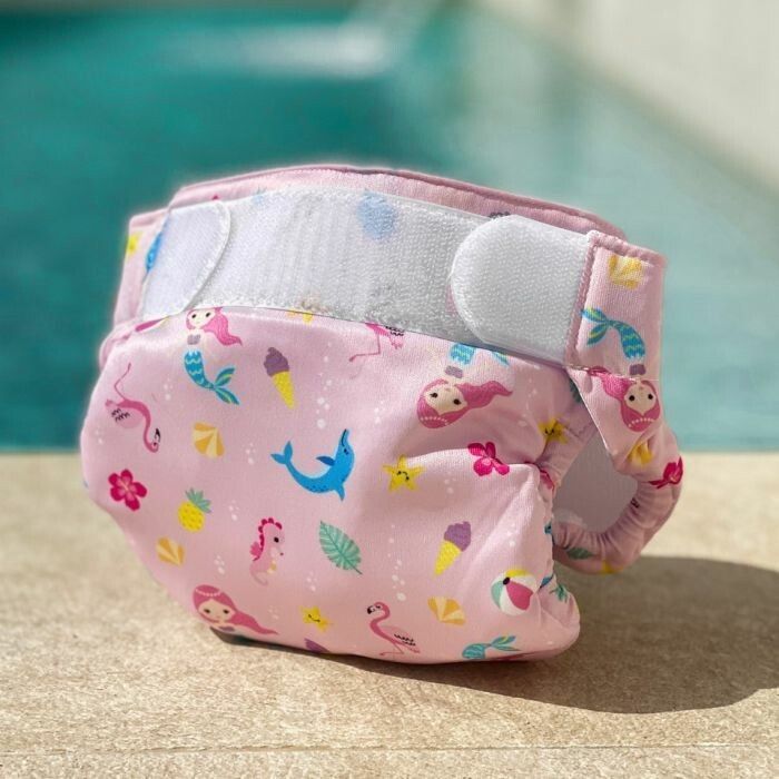 Pannolino da bagno rosa con motivi di sirene ed estivi. Chiusure in velcro. Appoggiato sul bordo piscina.
