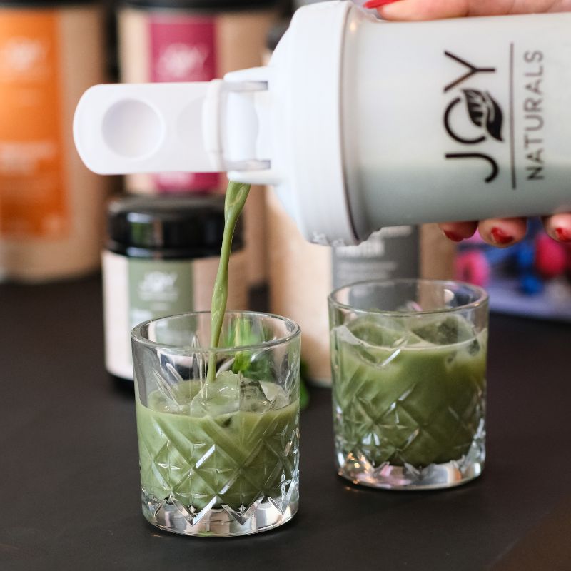 Bevanda verde versata da uno shaker in due bicchieri. Bicchieri con cubetti di ghiaccio. Altri prodotti e shaker sullo sfondo.