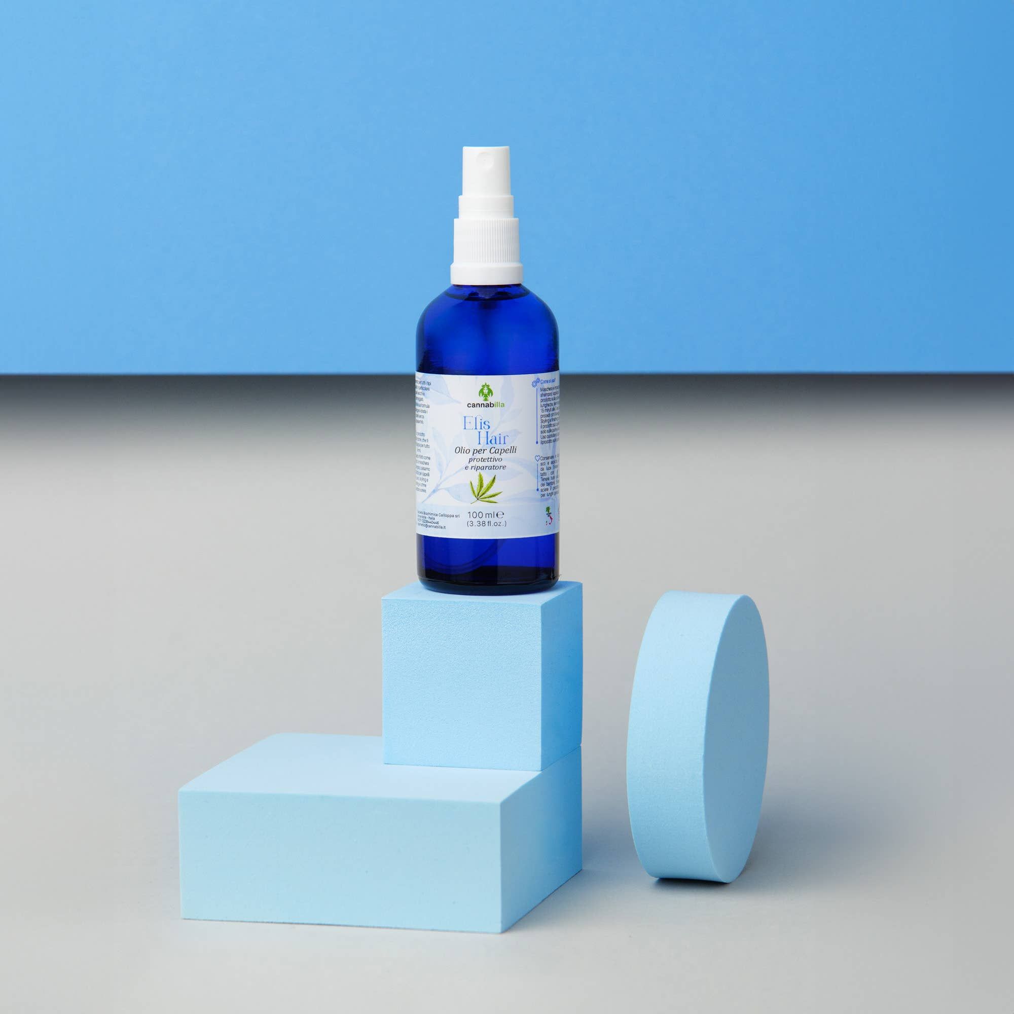 Flacone blu con vaporizzatore su piedistalli blu. Etichetta con 'Elis Hair' e informazioni prodotto. Sfondo blu e grigio.