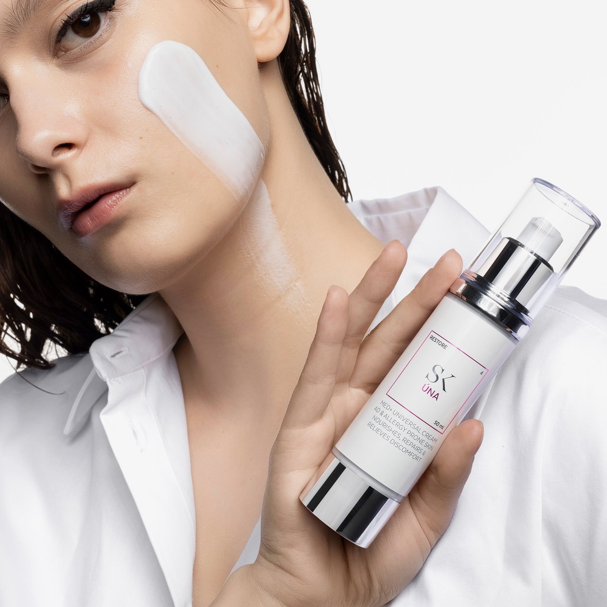 Donna che tiene un dispenser di crema. Crema sulla guancia. Testo: SK Úna, MED+ UNIVERSAL CREAM, per pelli allergiche.
