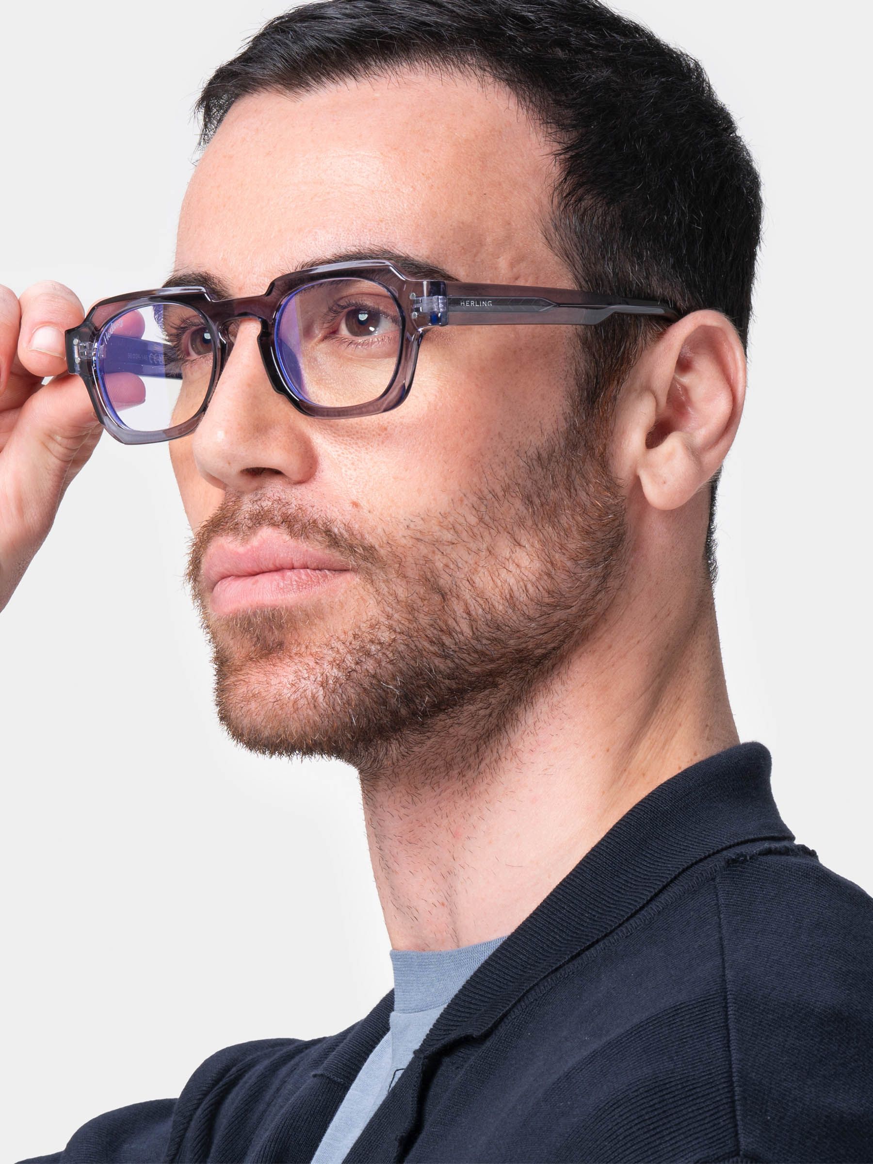 Uomo con occhiali. Montatura grigio-viola, lenti rettangolari. Ponte nasale nero. Logo Herling sull'asta. Camicia blu scuro.