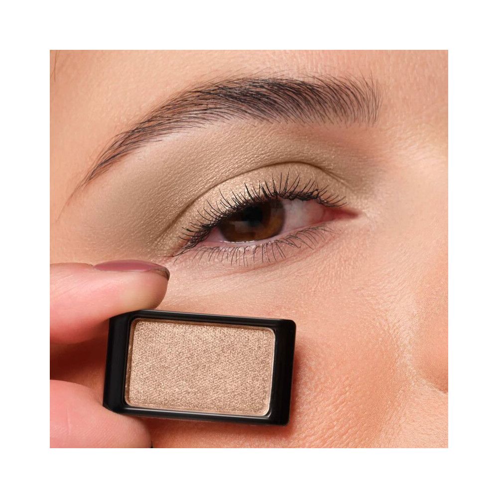 Un ombretto singolo in un contenitore nero. L'ombretto è marrone chiaro e viene tenuto davanti a un occhio.