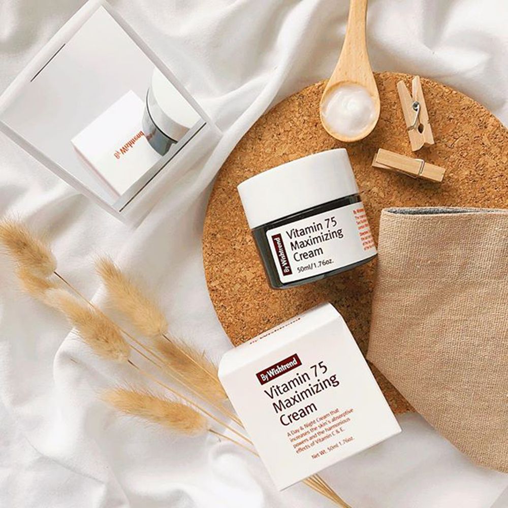 Prodotto su una tavola di sughero con accessori. Vaso e scatola con testo rosso: Vitamin 75 Maximizing Cream. Marchio: By Wishtrend. Su tessuto bianco.