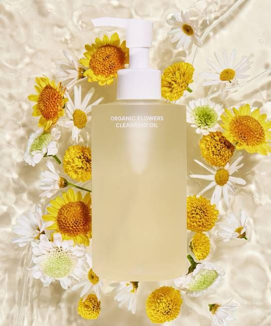 Flacone trasparente con erogatore bianco, circondato da fiori. Scritta: ORGANIC FLOWERS CLEANSING OIL.