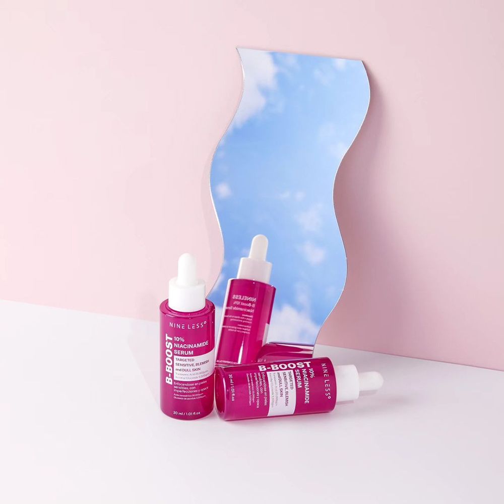 Tre flaconi di siero rosa davanti a uno specchio. Scritta: NINE LESS, B-Boost, 10% Niacinamide Serum. Lo specchio mostra il cielo blu.