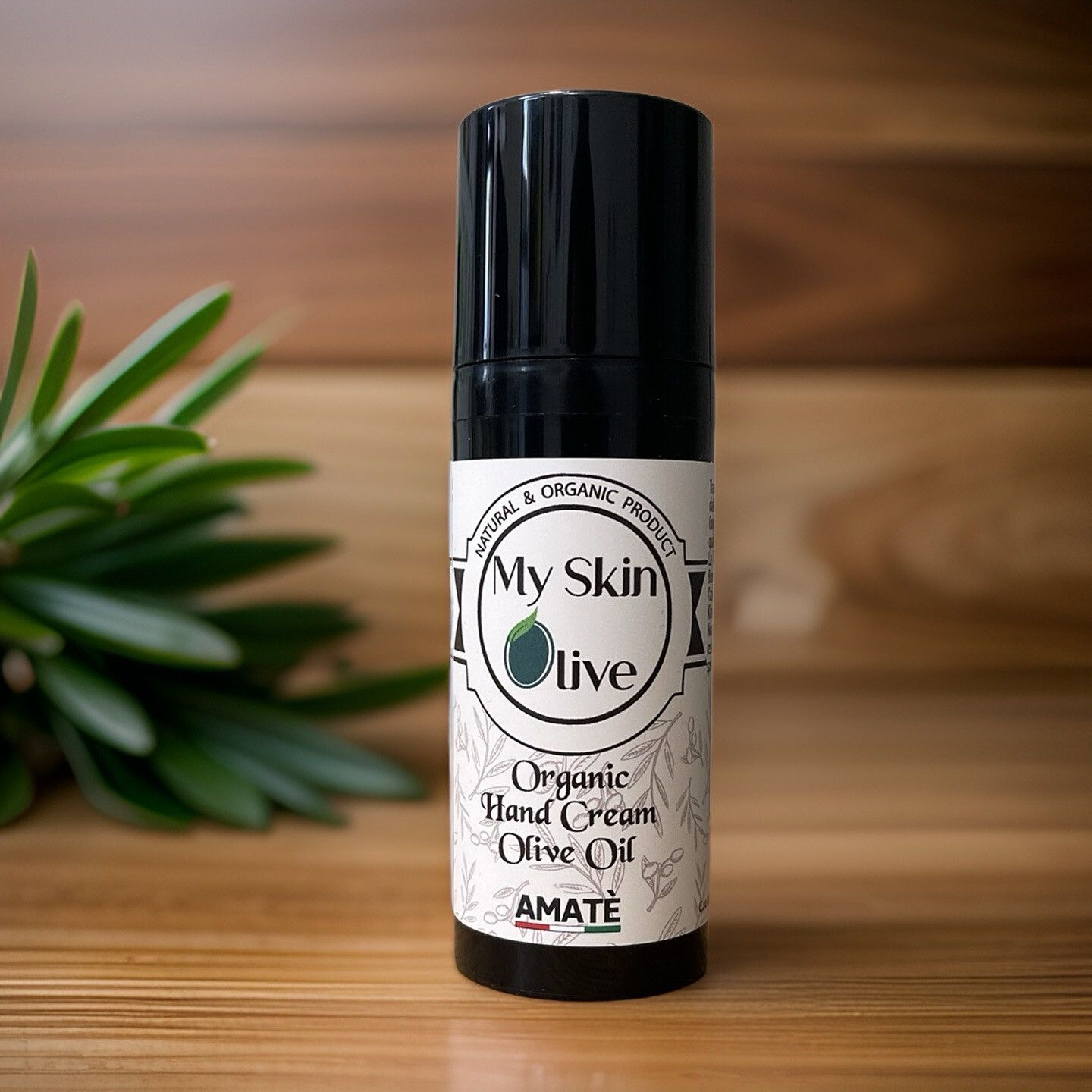 Flacone nero davanti a legno e foglie. Scritta: My Skin Olive, Organic Hand Cream Olive Oil, Amatè.