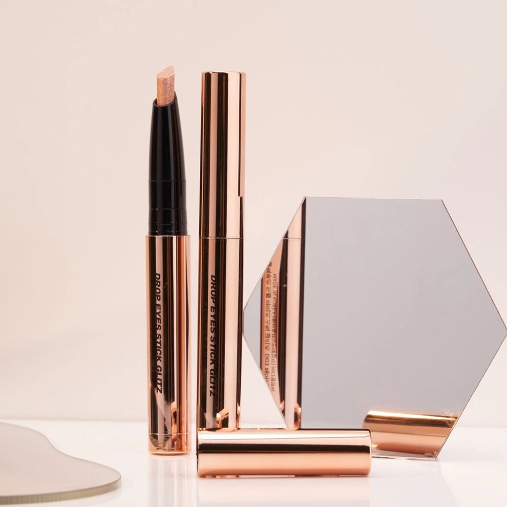 Tre ombretti Drop Eyes Stick Glitz rosa metallizzato, uno aperto, con specchio. Scritta: Drop Eyes Stick Glitz.