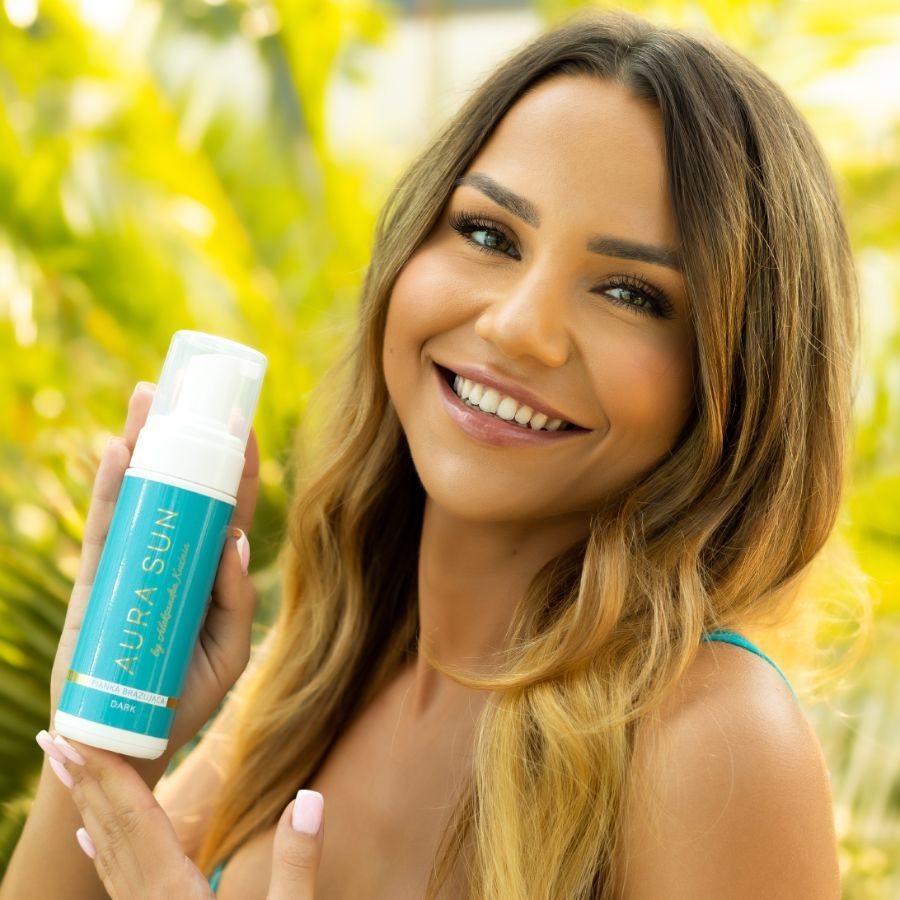 Una donna tiene in mano una bottiglia di mousse autoabbronzante AURA SUN. La bottiglia è turchese con tappo bianco.