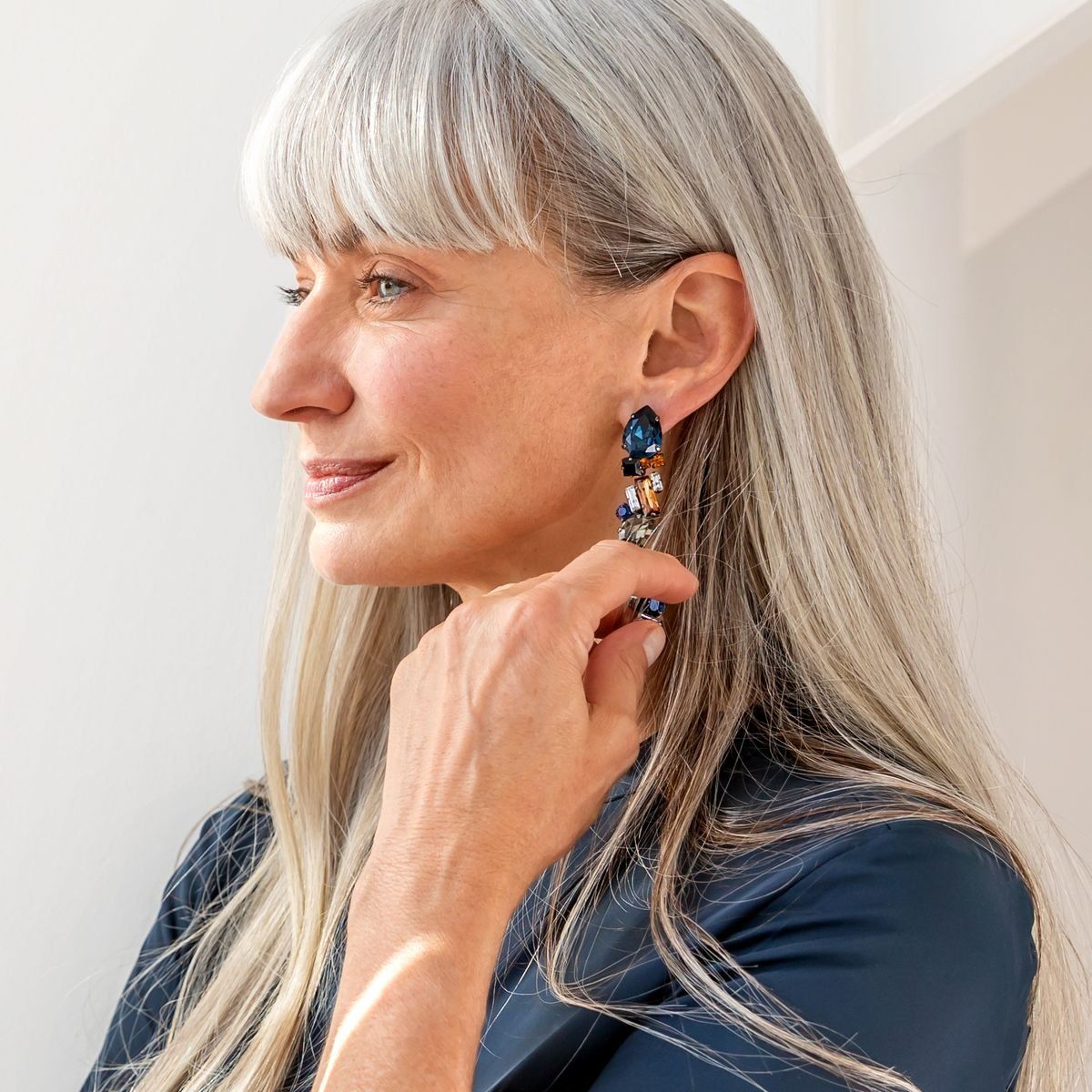 Donna con lunghi capelli grigi e orecchini. Si tocca l'orecchio. Gli orecchini sono blu e arancioni.