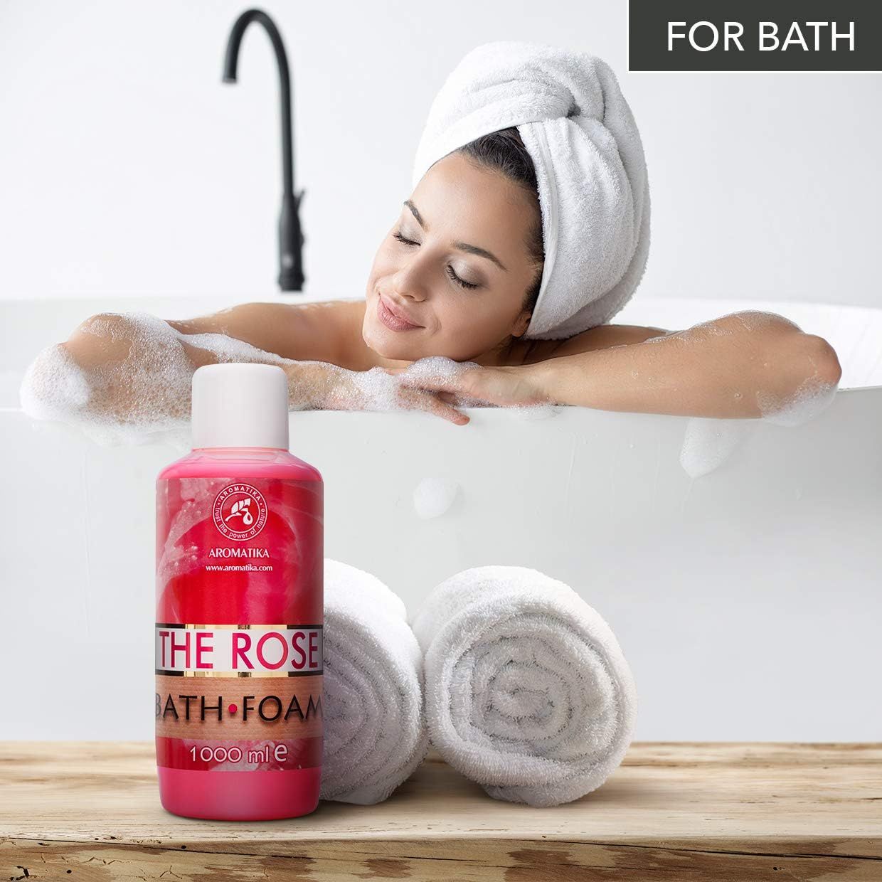 Flacone rosa AROMATIKA THE ROSE BATH FOAM accanto a una vasca da bagno. Donna con asciugamano in testa si rilassa nel bagno.