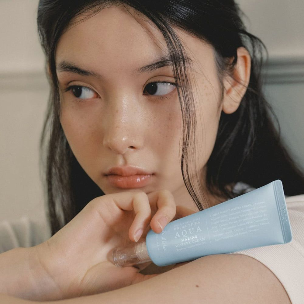 Una donna tiene in mano un tubo azzurro chiaro. Nome prodotto: Aqua Marine Watery Cream. Marchio: Dr. Althea. Applicatore trasparente.