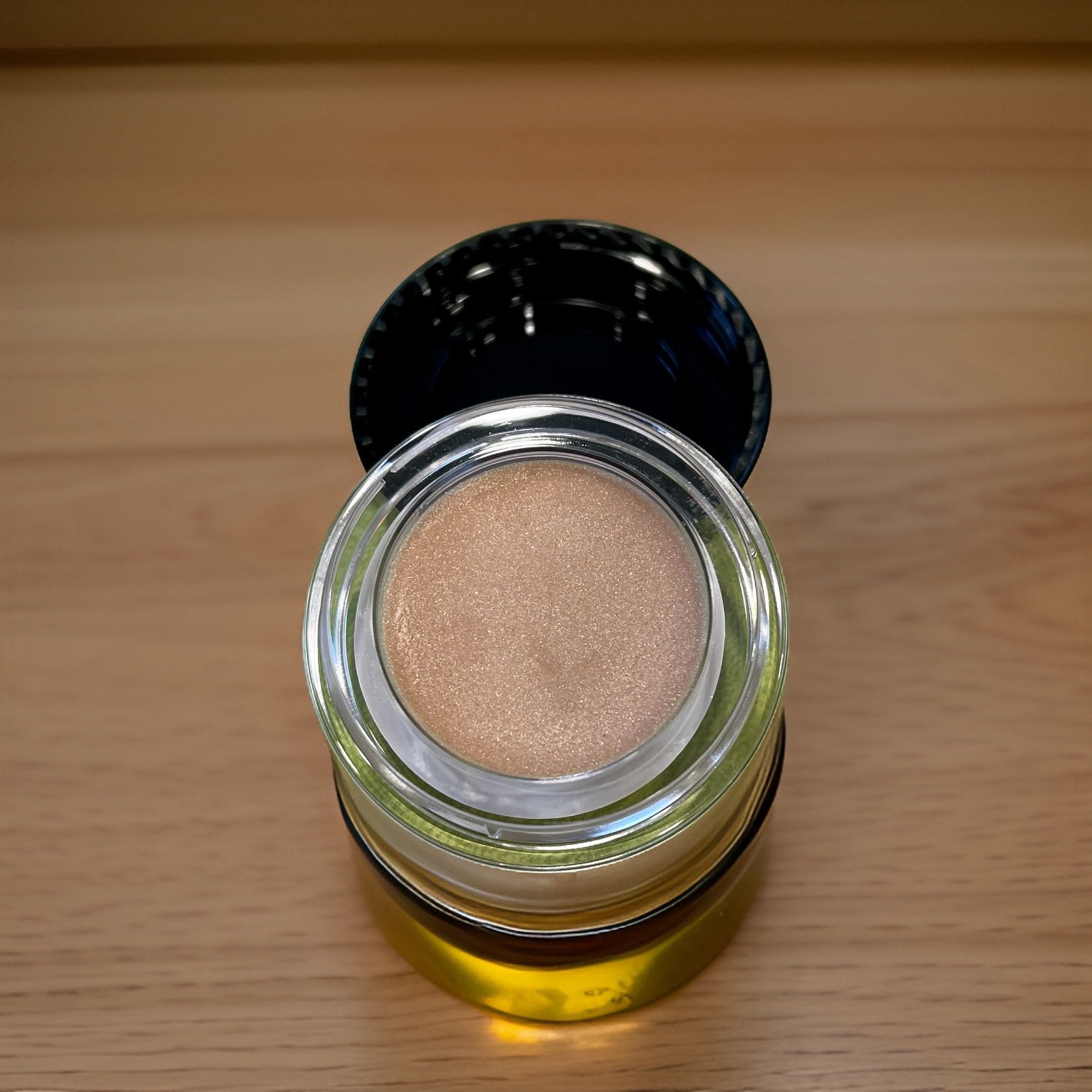 Vasetto di Sensual Gold Lip Balm Treatment, aperto. Coperchio accanto. Balsamo dorato nel vasetto, su fondo in legno.