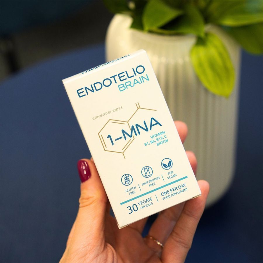 Mano che tiene una confezione del prodotto. Scritta: ENDOTELIO BRAIN, 1-MNA, 30 capsule vegane. Senza glutine, senza proteine del latte, vegano.
