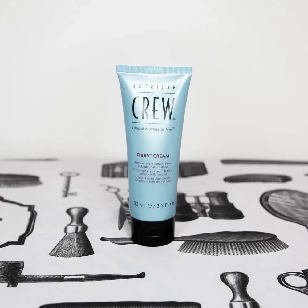 Tubo blu chiaro con tappo nero. Scritta: American Crew, Fiber Cream. Su tavolo con motivi da parrucchiere.