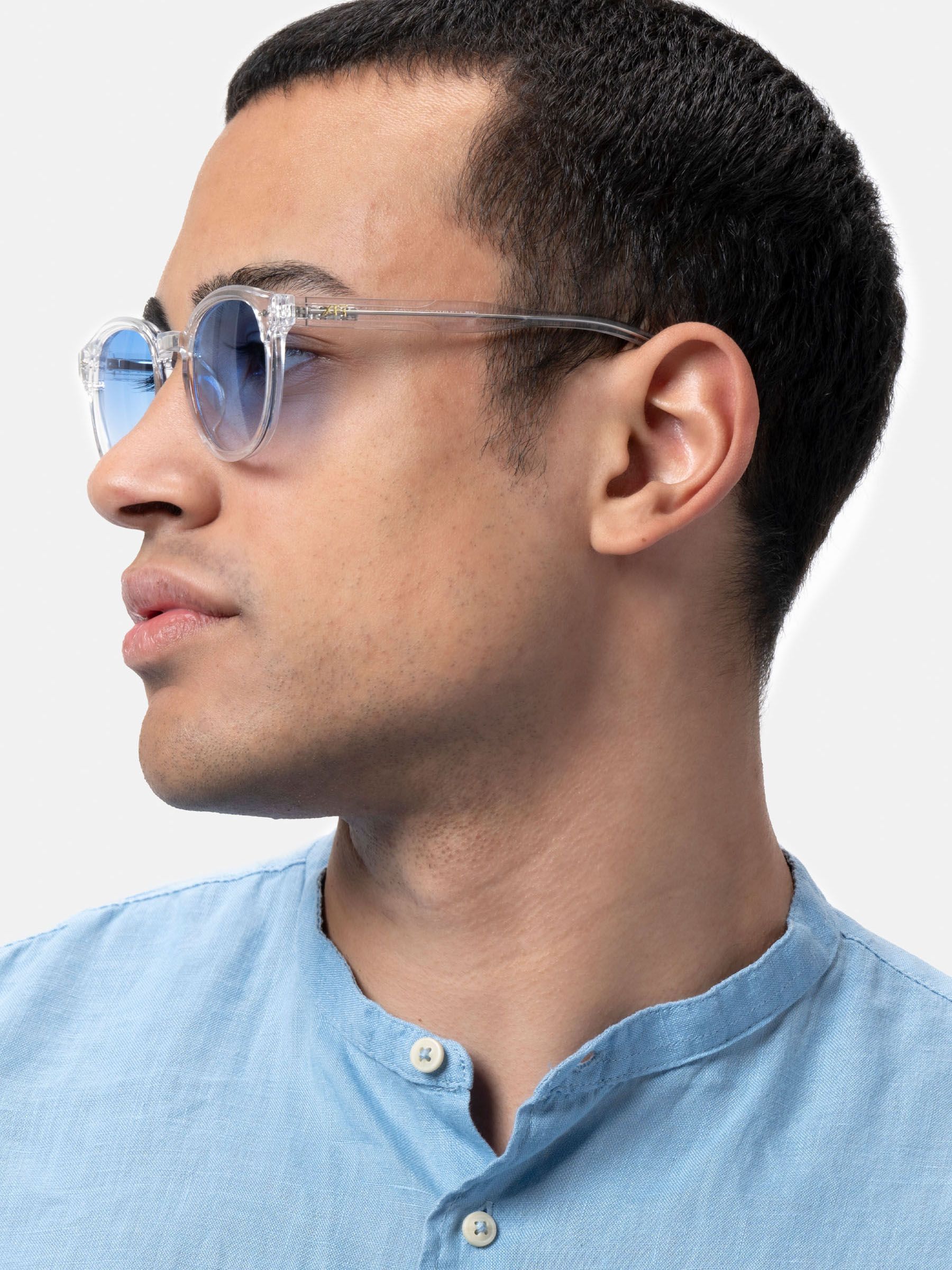 Uomo con occhiali da sole con montatura trasparente e lenti blu. Vista laterale, camicia azzurra.