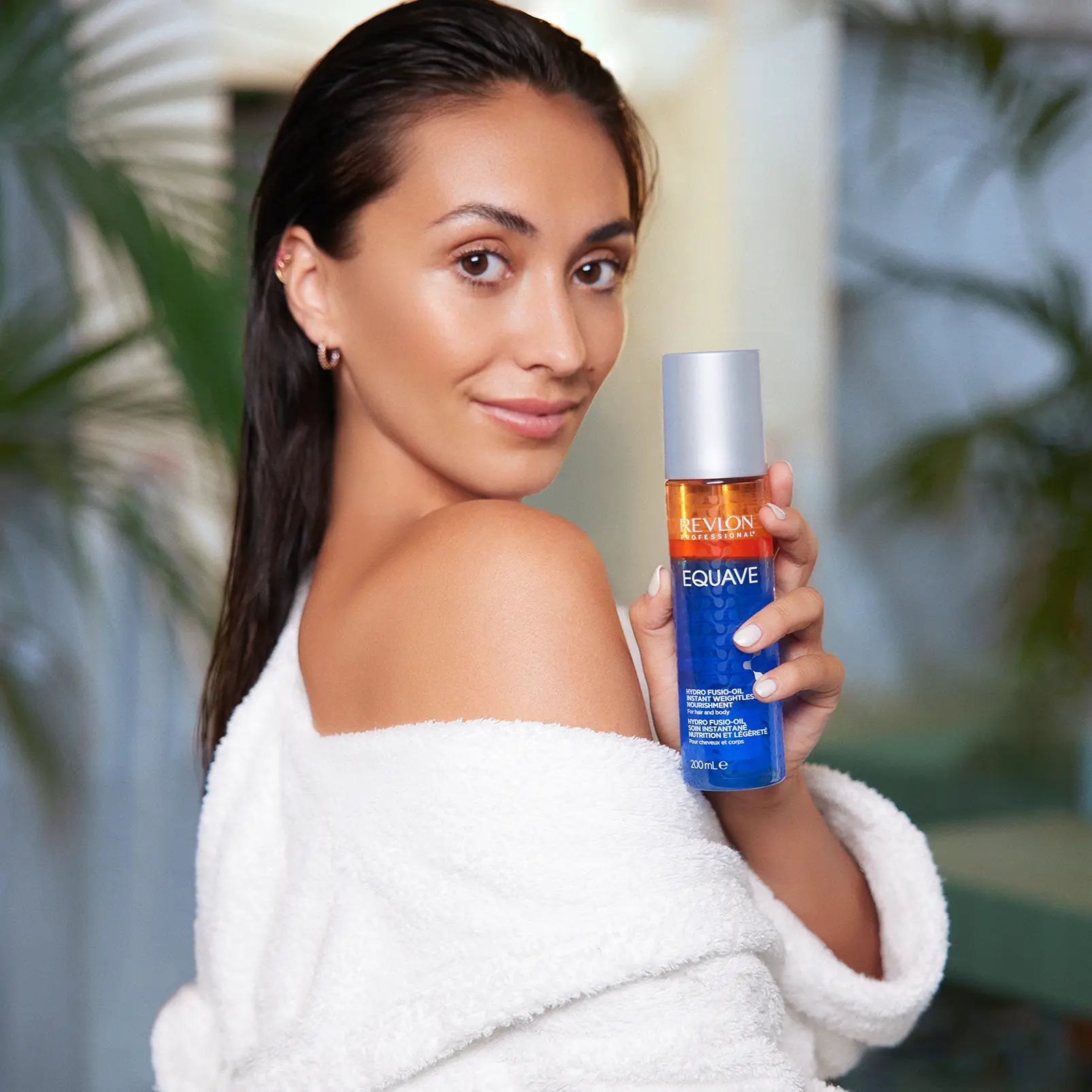 Donna in accappatoio tiene in mano un flacone. Flacone con tappo argentato, stampa arancione e blu. Nome del prodotto e marchio visibili.