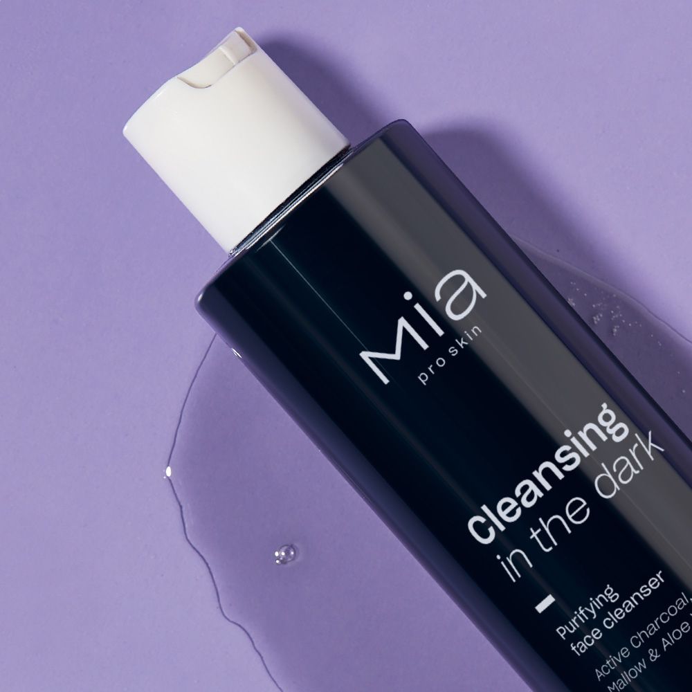 Flacone nero con tappo bianco. Scritta: Mia pro skin, Cleansing in the dark, detergente viso al carbone attivo. Su sfondo viola.