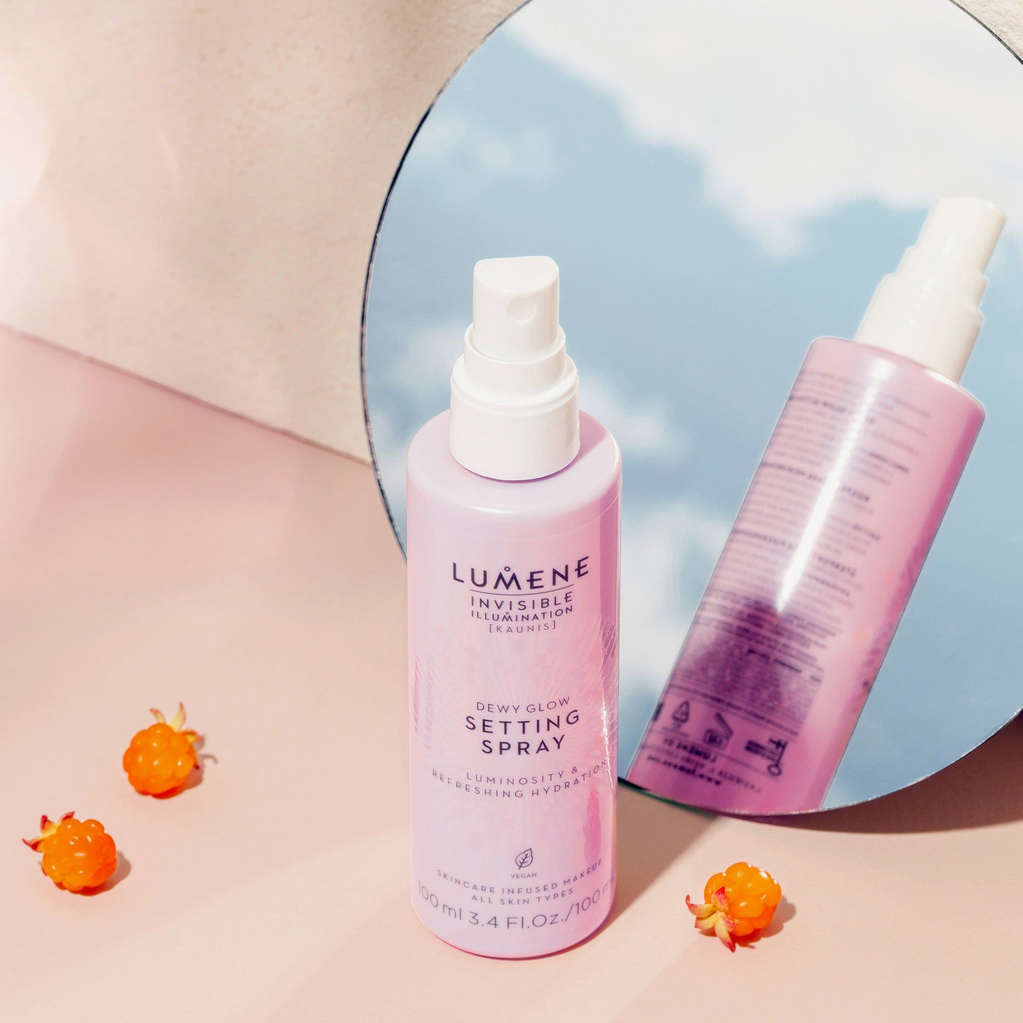 Due flaconi rosa con vaporizzatore bianco. Scritta: Lumene INVISIBLE ILLUMINATION Dewy Glow Setting Spray. Riflesso nello specchio. Bacche.