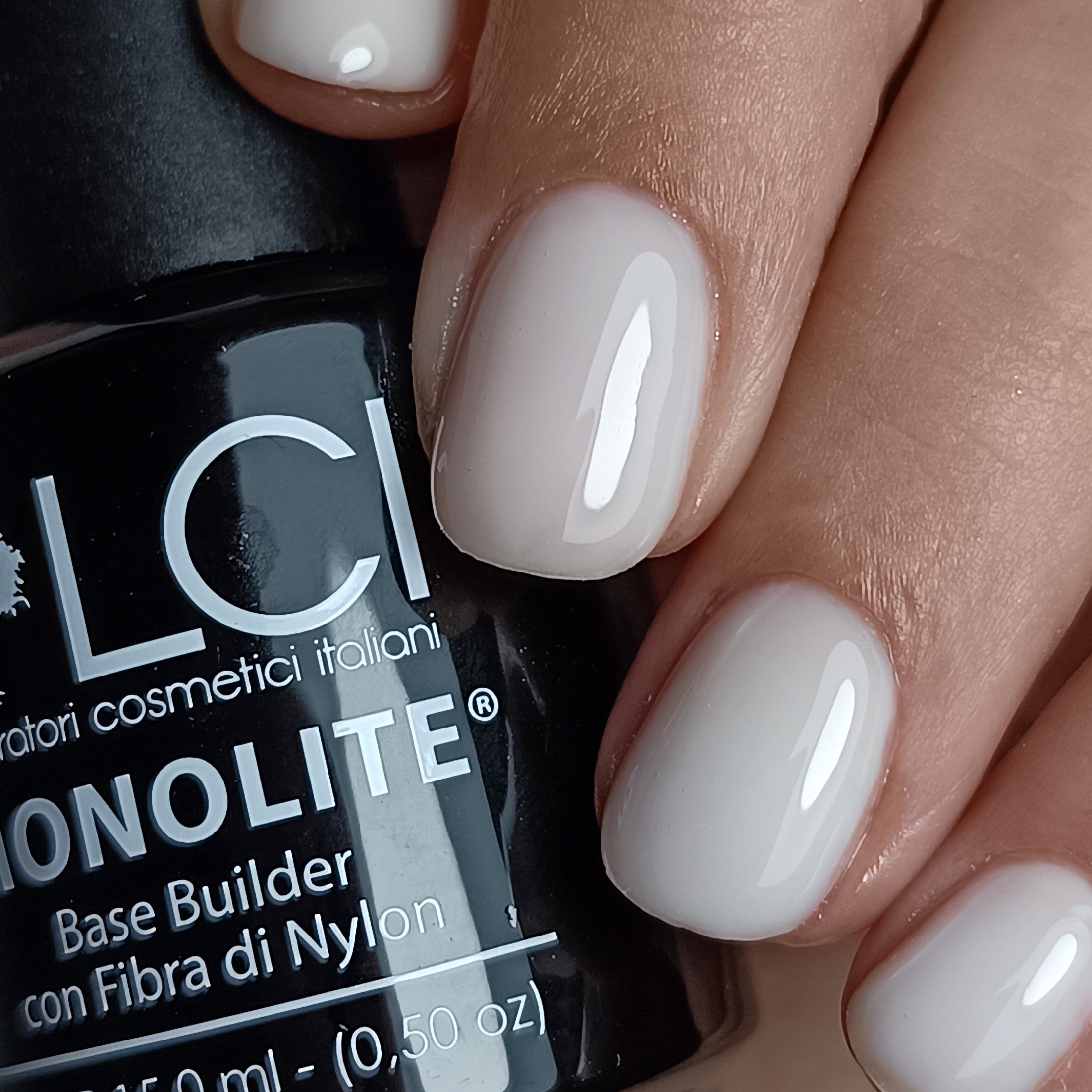 Primo piano di unghie con rivestimento in gel bianco. Sullo sfondo, un flacone con testo: LCI, MONOLITE, Base Builder.