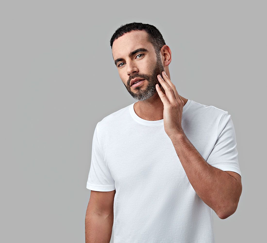 Un uomo con la barba si tocca il viso. Indossa una maglietta bianca.